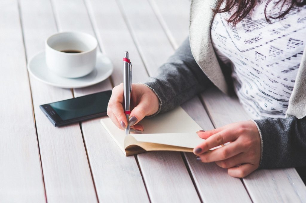 A person writing in a notebook at a table with a cup of coffee