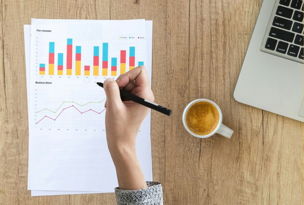 An overhead view of a hand annotating a printed bar-and-line chart beside a coffee and a laptop