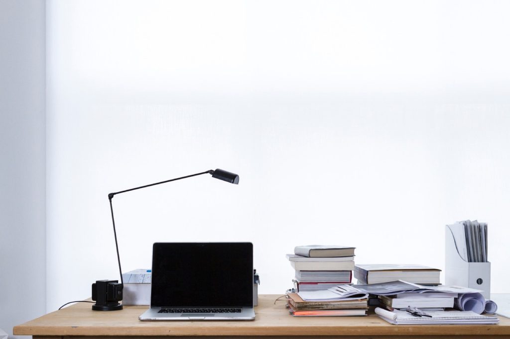 A tidy desk with a laptop, a lamp and a stack of files against a plain wall