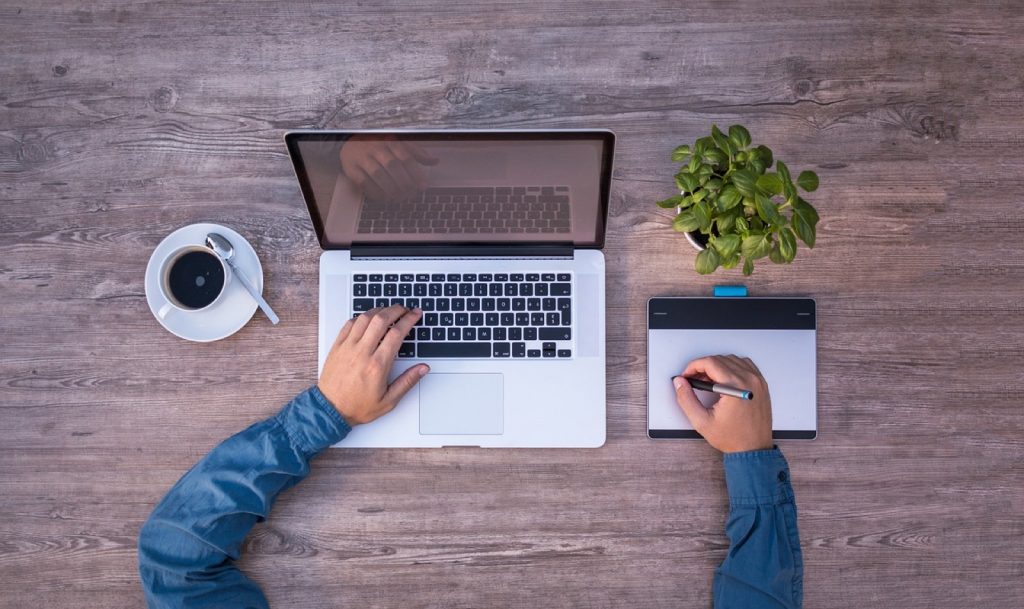 A laptop, a notepad and a cup of coffee on a wooden desk seen from above as someone works through numbers