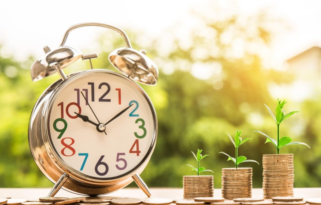 An alarm clock beside rising stacks of coins, suggesting time and cost