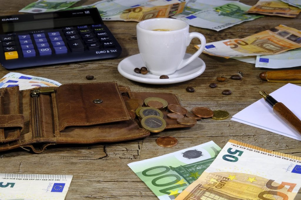 A leather wallet, scattered cash and coins, a calculator and a coffee on a wooden desk