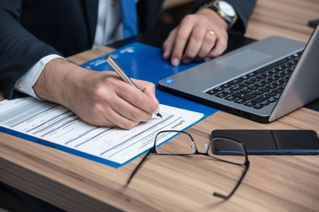 A person signing a printed agreement at a desk, with a laptop and reading glasses nearby