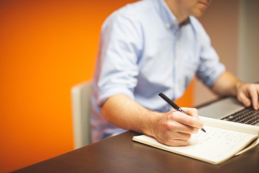 A person in a shirt writing in a notebook beside a laptop at a desk